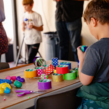 A young child at a table covered in colorful rolls of tape. The photo is taken from behind as the child looks down at the rolls. In the background are a few adults and other children.