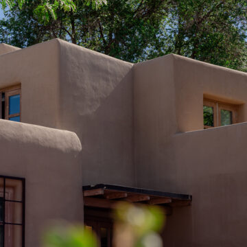 Exterior of an adobe building with two levels and several windows. The adobe building is framed by green trees and plants in the foreground and background.