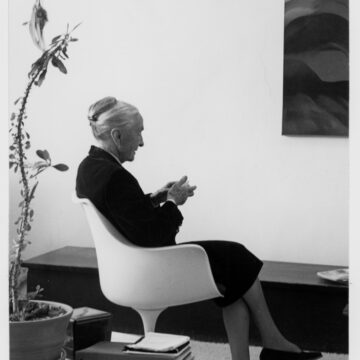 Full length profile view of O'Keeffe sitting in a white modular chair in her studio, her hair is pulled back into a bun, she wears a black suit coat and skirt, nylons, and black flat leather designer shoes while holding an object in her hand, hanging on the wall in viewer right is portion of one of her paintings (Black Place?), behind her to the left on a glass table are binders, books and a large spindly plant