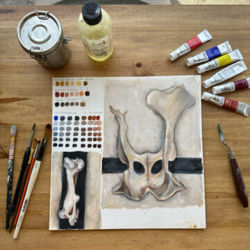 View from above of a painting of an animal pelvis bone and thigh bone on black strips. The paintings seem inspired from O'Keeffe's paintings of bones. The canvas rests on a wooden surface and surrounding it are art supplies including brushes, paints, oil thinner and a palette knife.