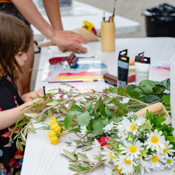 Photograph of a young child making an art piece on a table that is covered in art supplies and flowers.