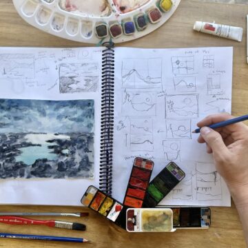 Open sketch book on table with watercolor paints, brushes, and pencils surrounding notebook. Notebook contains drawings