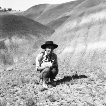 A black and white photograph of Georgia O'Keeffe. She wears a black pilgrim hat and casual denim clothes. The background there are flowing sandy hills.