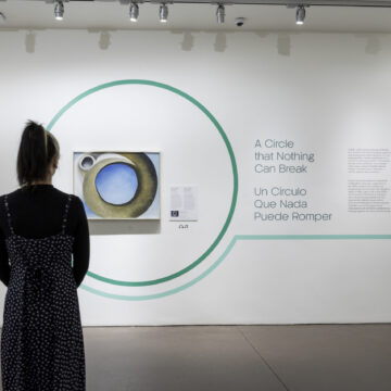 A person wearing a dress and their hair up stands in front of a painting of circular ram's horn on a gallery wall. On the wall is also a panel with text, a circular motif and the title 'A Circle that Nothing Can Break'
