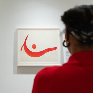 Photograph of a person with dark hair in buns wearing a bright red sweater from behind as they look at a painting by O’Keeffe on a white gallery wall. The painting shows a wave-like line with a dot above in shades of red.