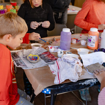 Photograph of a group of young children painting clay bowls made by hand at a table covered with art supplies.