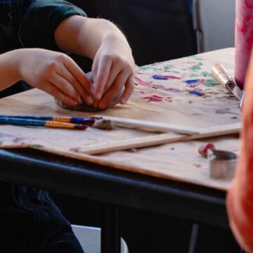Close-up photograph of a child's hands as they mold clay on a table. On the table are also some paint brushes.