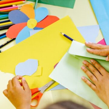 Photograph of the hands of children as they cut and fold colorful pieces of paper on a table strewn with pencils and other art supplies.