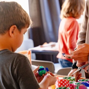Photograph of a child using colorful tape during an art activities. An adults hands approach from the right side of the frame.