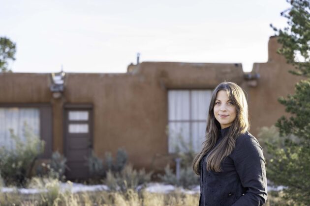 Photograph of a person with long brown hair and a dark jacket standing in front of an adobe home smiling at the camera. To the right of the frame is a tree.