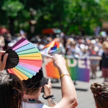 Photograph of hands waving a rainbow fan and a Pride flag in front of a crowd of people celebrating Pride. Behind are green trees.