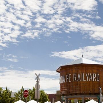 Photograph of a water tower with a silver roof and the writing 'Santa Fe Railyard' in silver lettering. Behind the tower is a blue sky and white clouds. Below are green trees and the top of little tents.