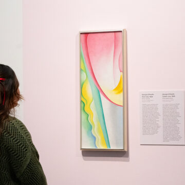 A person with long dark hair pulled up in small red bows looking at an abstract painting of a pink tulip. The painting is a vertical narrow canvas of an abstract section of a pink tulip. The upper quadrant of the picture plane encapsulates a large rounded draping petal shape in gradient shades of pink with an arc of yellow at the bottom of the form.