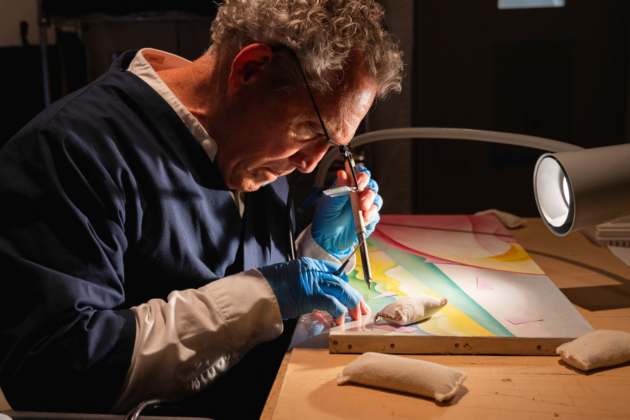 Photograph of a person wearing special eyeglasses and gloves working with specialized tools on a painting set on a table under bright lights.