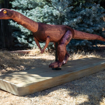 Photograph of a wooden model of a small proto-dinosaur, Effigia okeeffeae, on a wooden platform. Behind the sculpture are trees.