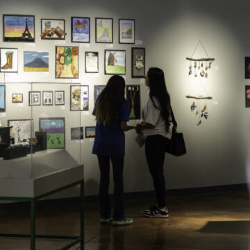 Two people stand in front of art made by students exhibited on a wall.