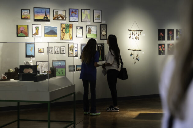 Two people stand in front of art made by students exhibited on a wall.