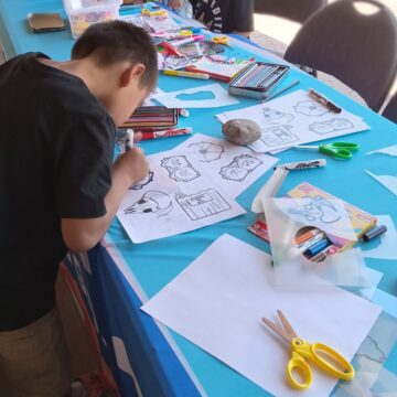 A young person with a dark shirt is color on a white piece of paper. They are bent over the table that is covered with art supplies. The table has a blue table-cloth.