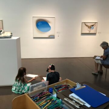 This photograph depicts the events at a First Friday art activity. Two children are sitting on the ground with clipboards. A box of art supplies and clipboards sits at the foreground of the image. To the right of the children a woman looks pensive as she sits on the bench and sketches.