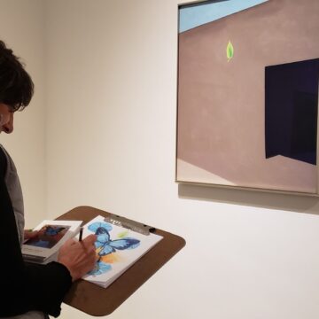 View of a person from the torso up holding a sketch book. They are looking at a painting of Georgia O'Keeffe's adobe patio.
