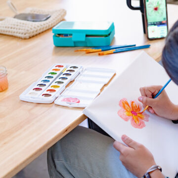 A young person with dark hair sits at a desk and paints a flower in shades of orange and red.