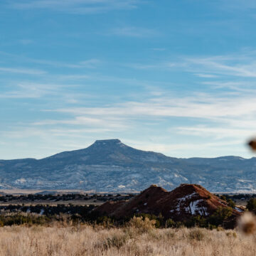 Photograph of Cerro Pedernal, or Tsee p'in, in the Tewa language. A flat-topped mountain with a vast desert landscape below. Behind the mountain is a blue sky and low clouds. To the right of the frame is a blurry plant.