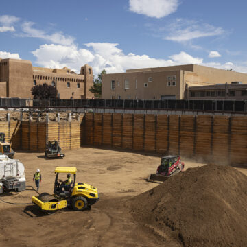 An excavation site with large wooden barriers, a dirt bottom, and construction trucks. Above the site are some adobe buildings and a blue sky.