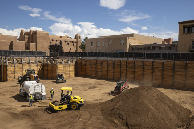 An excavation site with large wooden barriers, a dirt bottom, and construction trucks. Above the site are some adobe buildings and a blue sky.