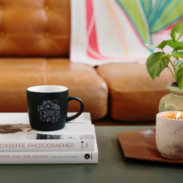 Photograph of a coffee table on which is a stack of books about Georgia O'Keeffe, a black mug with floral designs, a candle with a small flame, and a plant in a pot. Behind is a leather couch on which is draped a blanket in shades of pink and green based abstractly on O'Keeffe's painting of a Pink Tulip.