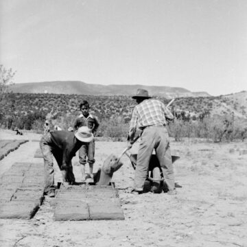 Black and white photograph of two people with a younger child using their hands and a shovel to make adobe bricks from a mud and straw mixture. Behind them is a desert landscape.