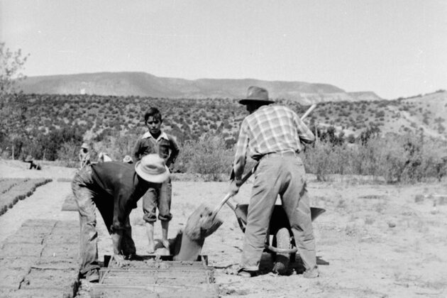 Black and white photograph of two people with a younger child using their hands and a shovel to make adobe bricks from a mud and straw mixture. Behind them is a desert landscape.