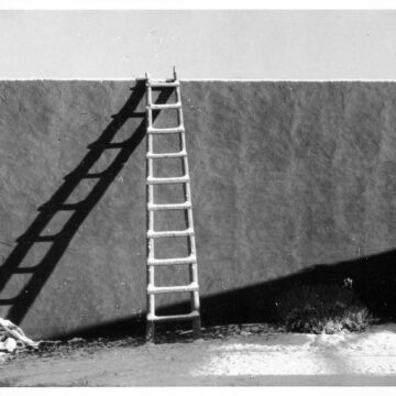 Black and white photograph of an adobe wall with a door on the left and a ladder leaning against the wall and casting a dark shadow against the wall. Below the ladder and along the bottom of the wall the ground is blanketed in white snow.