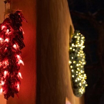 An adobe wall on which is a red ristra and a green wreath. Both are covered in lights. Behind the building is a dark sky.