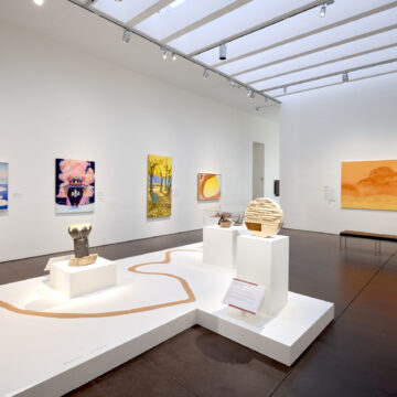 Photograph of a large gallery with white walls. Various colorful paintings adorn the walls and at the center of the room is a platform with objects including a drum and a rock sculpture.