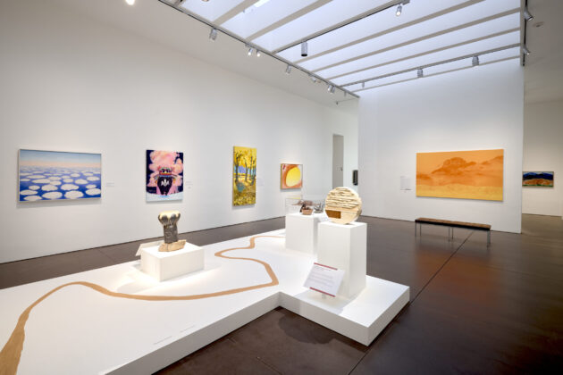 Photograph of a large gallery with white walls. Various colorful paintings adorn the walls and at the center of the room is a platform with objects including a drum and a rock sculpture.