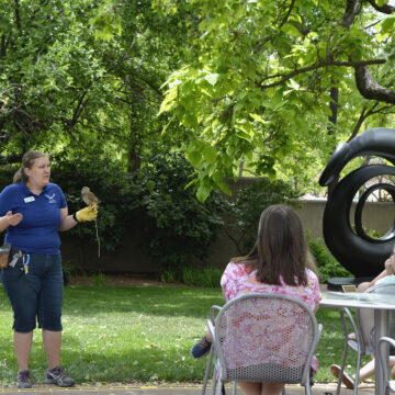 A person holding a small owl in front of a group of people sitting at tables outdoors. Behind the owl handler are green trees and an abstract sculpture