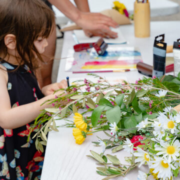 Photograph of a young child making an art piece on a table that is covered in art supplies and flowers.