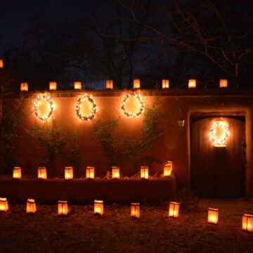 A wall with a wooden doorway where farolitos light up the darkness. Along the wall are also some wreaths wrapped in bright lights.