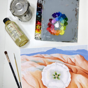 An image from above of a small canvas where a white flower sits on a desert background. Above the small painting is a palette with colorful paints and paint tubes.