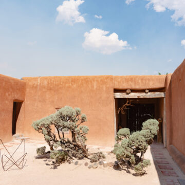 An inner courtyard with a blue sky above. In the courtyard are a few small green bushes, the frame of a wire butterfly chair and a covered well.