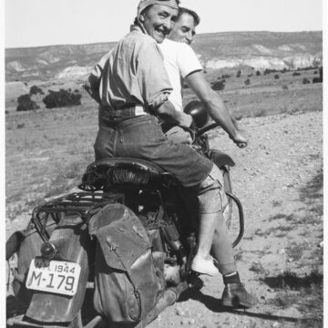 Georgia O'Keeffe on the back of a motorcycle holding on to a male friend (Maurice Grosser) who is driving. She is turning back looking towards the camera, in an expansive New Mexican landscape. She is wearing denim jeans, a chambray shirt, a cap with goggles, and sneakers.