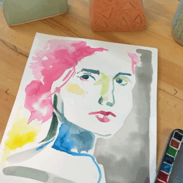 A watercolor portrait with colorful paints rests on a wooden table. At the bottom of the photograph a colorful palette is visible.