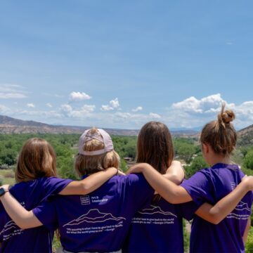 A group of four young people stand with their arms on each other's shoulders looking out over a desert landscape. All four wear matching purple t-shirts.