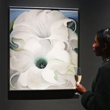 Photograph of a person with their hair tied up standing in front of a large painting of white flowers against a dark wall. In their hand they hold a filled glass.