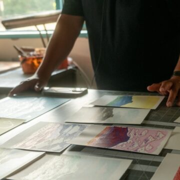 A photograph of a person's hands hovering over several small paintings of a flat-topped mountain laid on a table.