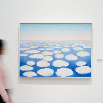 Photograph of a large painting by Georgia O'Keeffe of some fluffy white clouds across a blue horizon. The painting hangs on the white walls of a museum gallery and in front, a person's blurry silhouette is visible as they walk in front.
