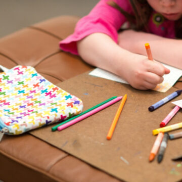 Photograph of a young child's arms and hands as they draw on a piece of paper with colorful pencils.