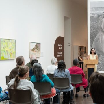 A person stands at a podium and address a crowd of people seated in a gallery with white walls.