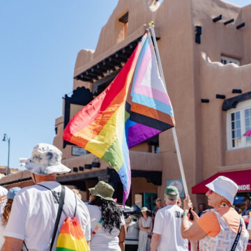 A group of people wearing white shirts with the Museum logo walk along a street in downtown Santa Fe. A person waves a large colorful Pride flag. Several people in the group have hats and colorful bags.