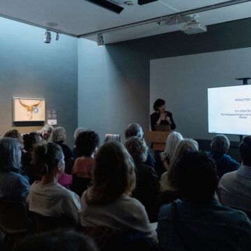 A person stands at a podium in a dark room with a screen to their left and a painting on the wall to their right. They speak to a large crowd of seated listeners.